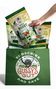 
                  
                    Hand holding a bag of Gussy's Gut Daily Boost supplements, surrounded by additional products in a green Gussy's Gut tote.
                  
                