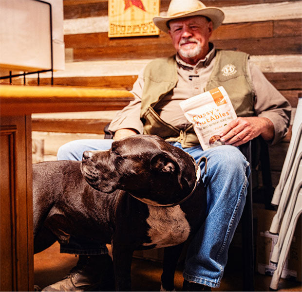 Man with a dog, holding organic fermented dog treats labeled "Cussy & Outables," in a cozy, rustic setting.